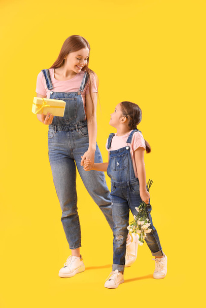 Cute little girl and her mother with gift holding hands on yellow background - Φωτογραφία, εικόνα