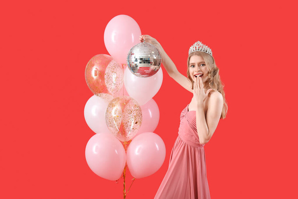 Удивлённая девушка-подросток в выпускном платье с диско шаром и воздушными шарами на красном фоне - Фото, изображение