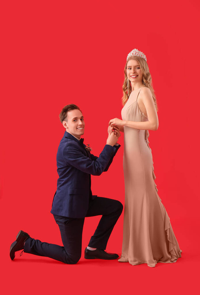 Young man standing on one knee in front of his prom date against red background - 写真・画像