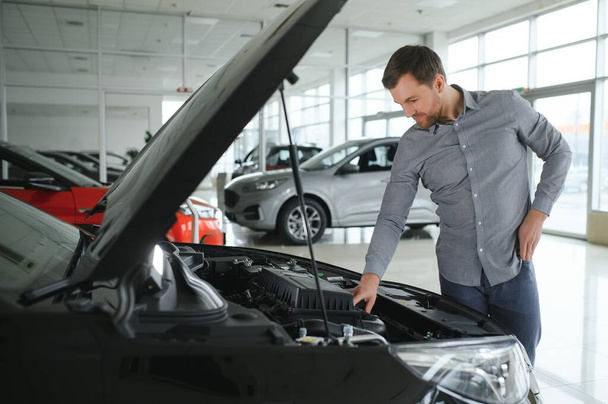 Looks under the hood of automobile. Young man in the car dealership - Foto, Imagen