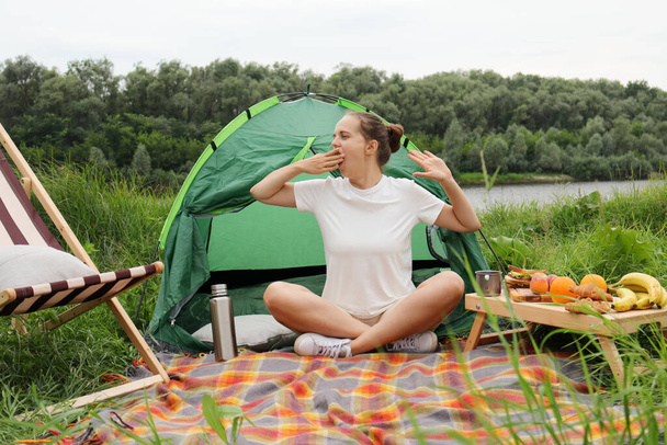 Photo of calm sleepy woman with bun hairstyle wearing white t shirt sitting near the tent near the river, waking up early, sitting with crossed legs, yawning, stretching arms. - Fotoğraf, Görsel