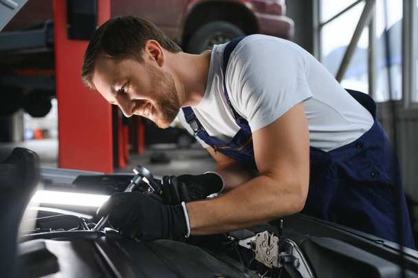 Handsome young male auto mechanic in special uniform clothes holding a flashlight, looking for breakdown and repairing under the hood in the car engine in a car workshop. - Fotoğraf, Görsel
