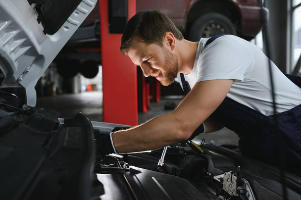Handsome young male auto mechanic in special uniform clothes holding a flashlight, looking for breakdown and repairing under the hood in the car engine in a car workshop. - Foto, Bild