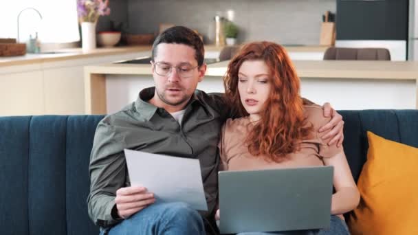 Couple sit on couch with laptop holding document pay bills, payment of the fine count expenses, make budget control, manage finances, review overdue loan payments feel stressed. Crisis, lack of money - Séquence, vidéo