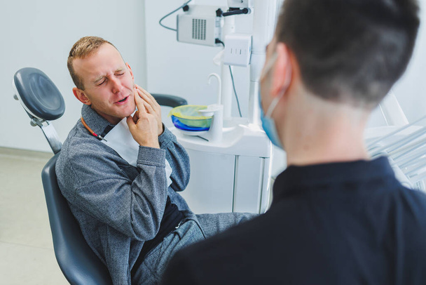 Молодой врач-дантист рассказывает пациенту-мужчине о стоматологической помощи. Мужчина сидел в стоматологическом кресле на приеме у врача. Современное лечение зубов - Фото, изображение