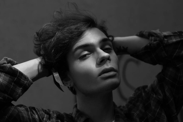 Closeup portrait of relaxed apathetic young guy in plaid shirt running hands through his dark frizzy hair posing in dark room. Black and white shot - Φωτογραφία, εικόνα
