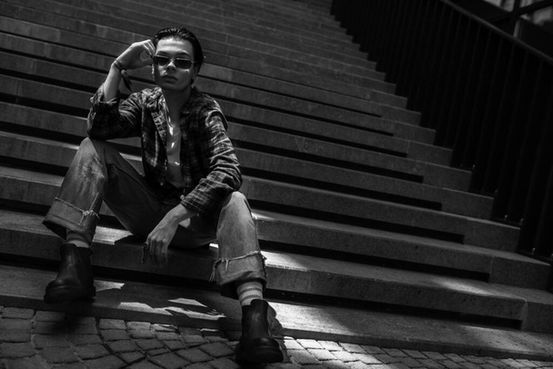 Уверенный в себе молодой модельный парень, позирующий на улице, сидящий на каменной лестнице, регулируя очки. Черно-белый портрет - Фото, изображение