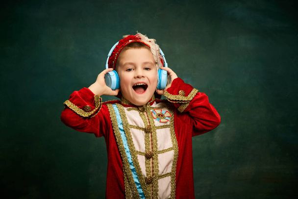 Cheerful little boy dressed up as medieval little prince and pageboy listening to music over dark vintage style background. Retro fashion, emotions, music concept. Kids looks happy - Photo, Image