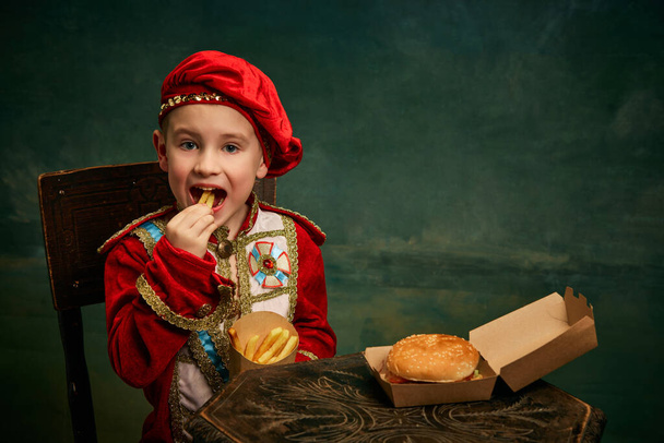 Happy cheerful little boy wearing costume of medieval page boy and prince eating hamburger with french fries over dark green background. Fast food, happiness, childhood and tastes concept - Photo, image