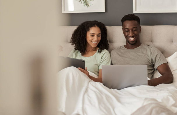 Smiling young multiracial couple sitting up in their bed at home in the morning and using a laptop and digital tablet - Fotó, kép