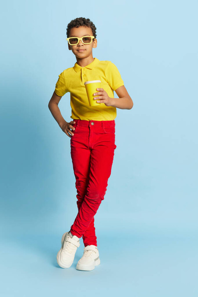 Koude limonade op warme dagen. Modieuze kleine Afrikaanse jongen in stijlvolle zonnebril en geel zomershirt en rode broek poserend over een blauwe achtergrond. Concept van mode, geluk, kinderen emoties - Foto, afbeelding