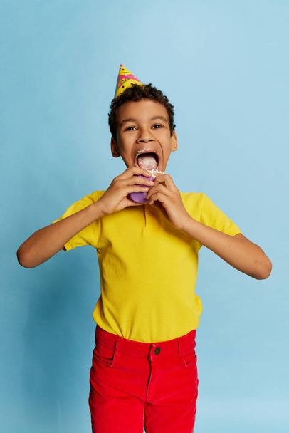 Delighted school age boy in bright colors clothes with birthday cone eating sweet cake isolated on blue background. Concept of fashion, art photography, style, holidays, dreams, ad. Happiness - Foto, imagen