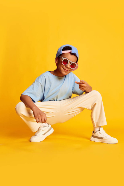 Portrait dynamique du petit garçon africain, danseur hip-hop dans des vêtements de rue élégants assis sur un fond jaune vif. Concept de musique, de bonheur dansant. Il a l'air heureux et sportif. - Photo, image