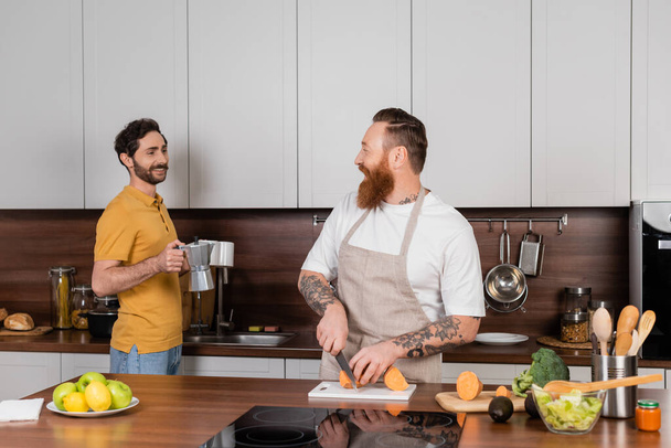 Smiling gay man holding coffee and cups near husband cooking in kitchen  - Foto, immagini