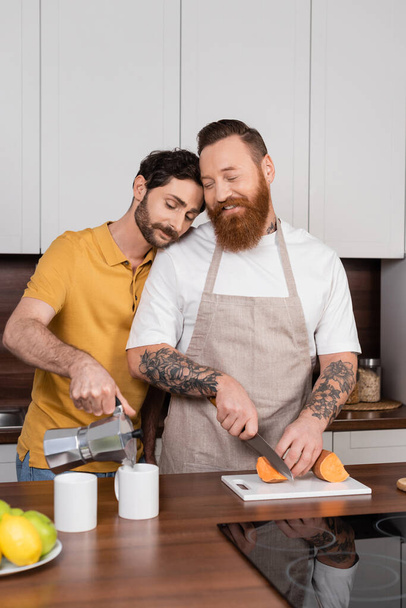 Homosexuell Mann gießt Kaffee in der Nähe lächelnden Ehemann schneiden Süßkartoffeln in Küche  - Foto, Bild
