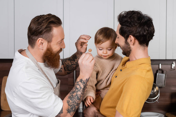 happy gay fathers holding daughter and barrette in kitchen  - Foto, Imagem