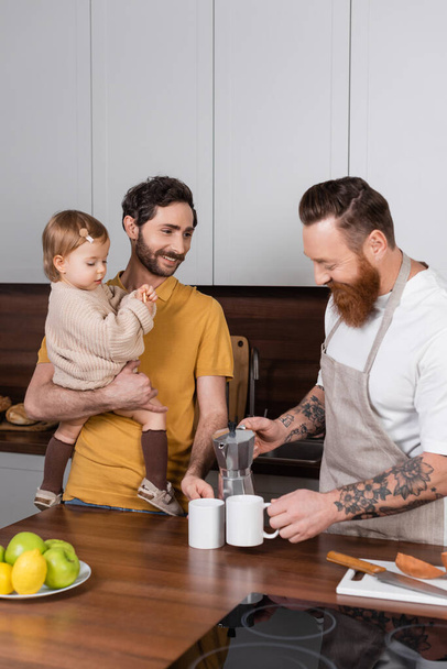 Sorrindo gay homem derramando café perto parceiro com filha no cozinha  - Foto, Imagem