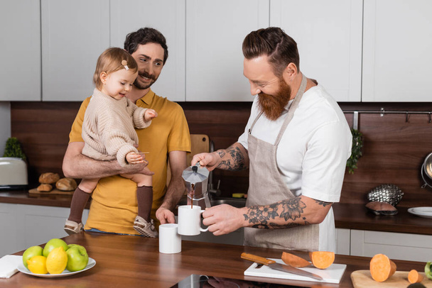 Positive tattooed gay man pouring coffee near partner holding daughter in kitchen  - Fotó, kép