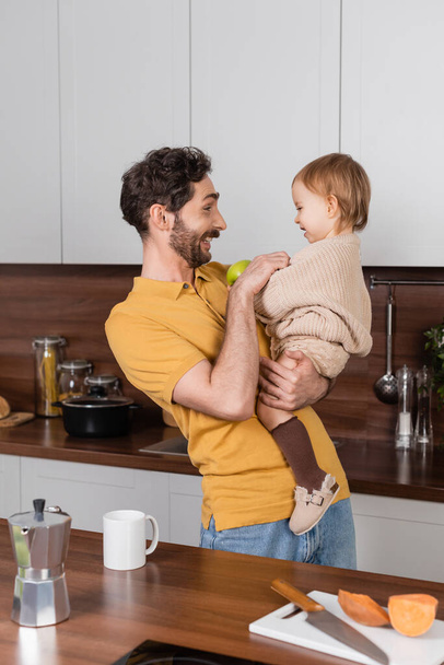 Vue latérale de l'homme souriant tenant fille tout-petit avec pomme dans la cuisine  - Photo, image