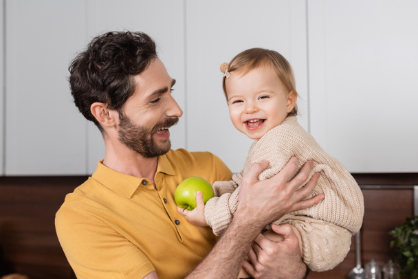 Smiling father holding toddler daughter with apple in kitchen  - Foto, imagen