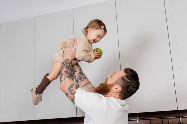 Tattooed man holding toddler daughter with apple in kitchen  - Фото, изображение