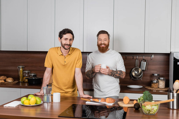 Smiling gay couple with coffee looking at camera in modern kitchen  - Zdjęcie, obraz