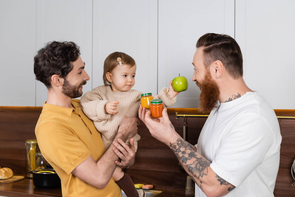 Gay couple holding toddler daughter and baby food in kitchen  - Foto, imagen