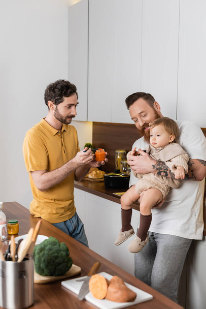 Χαμογελαστός ομοφυλόφιλος άντρας που κρατάει την κόρη του μικρού παιδιού κοντά στον σύζυγο με βρεφικές τροφές στην κουζίνα  - Φωτογραφία, εικόνα