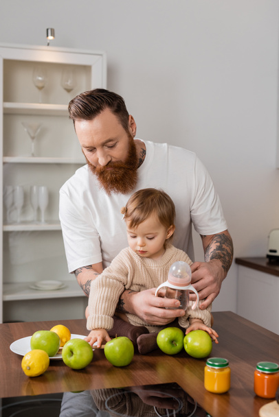 Tattooed dad holding baby bottle near daughter and fruits in kitchen  - Fotoğraf, Görsel