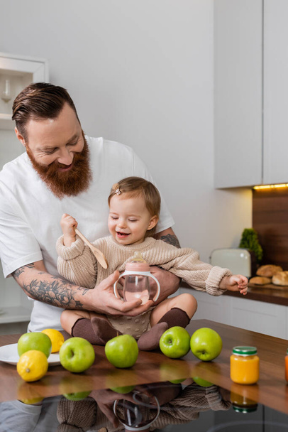 Bearded man holding baby bottle near daughter with spoon and fruits in kitchen  - Foto, Imagem