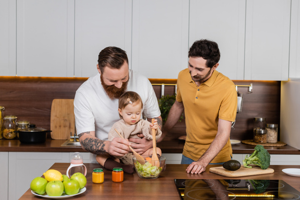 Gay man cooking salad near toddler daughter and partner in kitchen  - Fotoğraf, Görsel