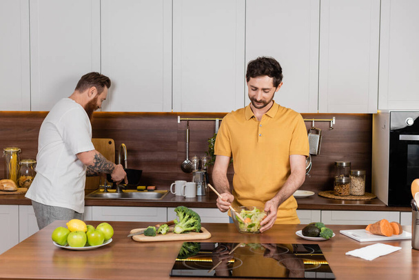 Gay man cooking salad near partner and food in kitchen  - Fotografie, Obrázek