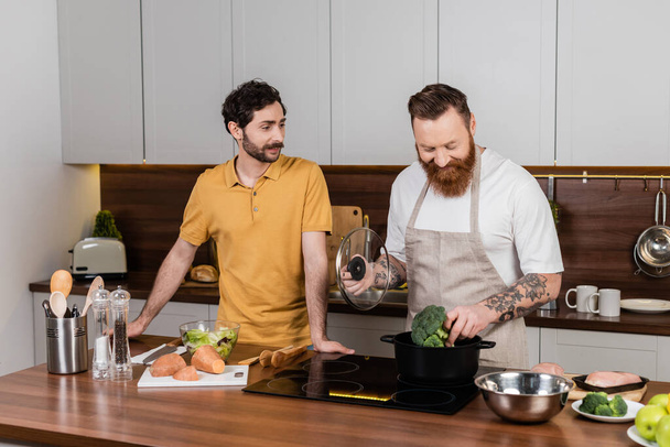 Tattooed gay man putting broccoli in pot while cooking near partner in kitchen  - Фото, зображення
