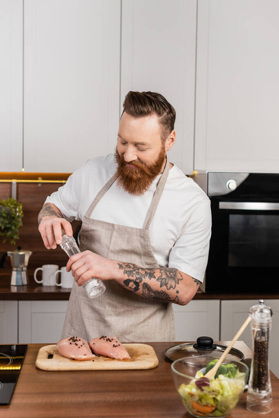 Bearded and tattooed man salting chicken fillet near fresh salad in kitchen  - Foto, Imagem