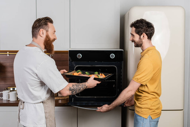 Vue latérale du couple gay mettant de la viande et des légumes sur une plaque de cuisson au four à la maison  - Photo, image