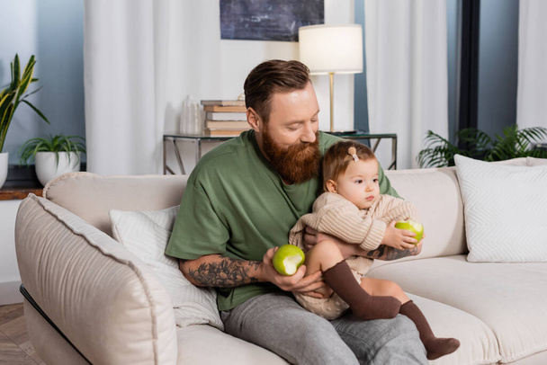 Bearded father holding apple and toddler daughter on couch at home  - Фото, изображение