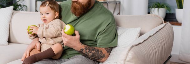 Tattooed father holding fresh apple and toddler daughter in living room, banner  - Φωτογραφία, εικόνα