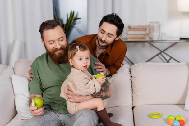 Smiling gay couple looking at baby daughter with apple on couch at home  - 写真・画像