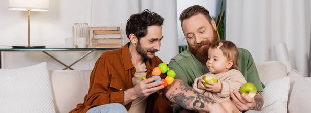 Smiling gay man holding toy near daughter and partner with apples, banner  - Фото, изображение
