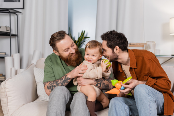 Happy gay couple holding baby girl with apple in living room  - Foto, Bild