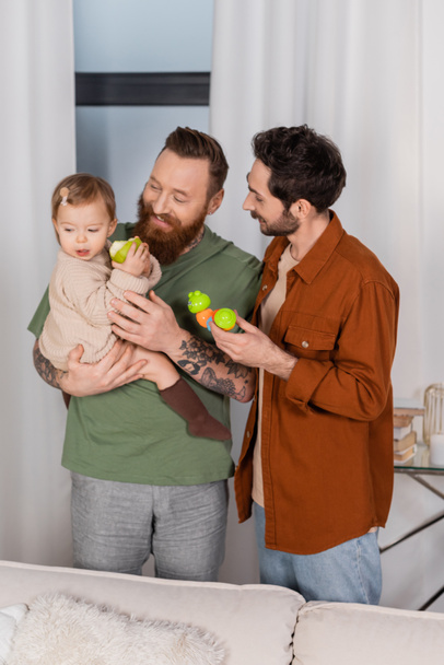Cheerful gay parents holding baby girl with apple and toy at home  - Foto, Imagem