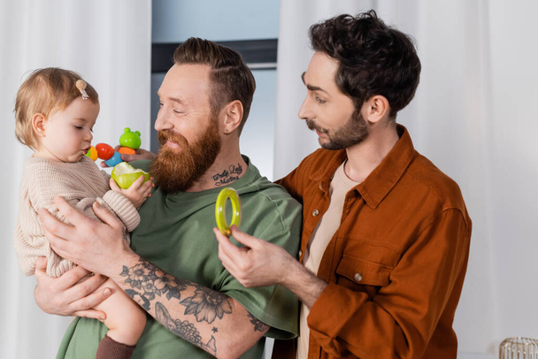 Smiling gay couple holding toys and baby girl with apple at home  - 写真・画像