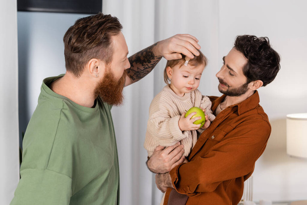 Mismo sexo padres sosteniendo bebé hija con manzana en sala de estar  - Foto, imagen