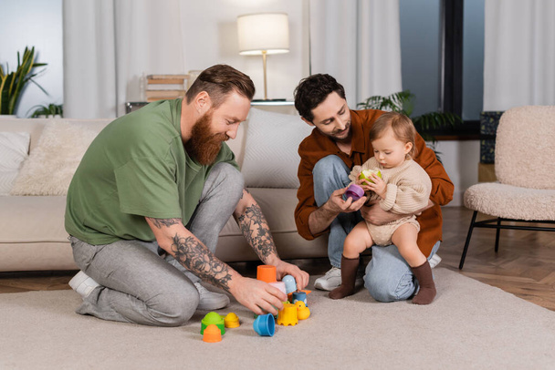 Same sex parents holding toys near baby girl with apple at home  - Foto, imagen