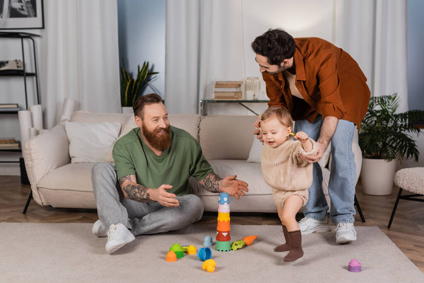 Cheerful homosexual parents playing with baby daughter in living room  - Фото, изображение