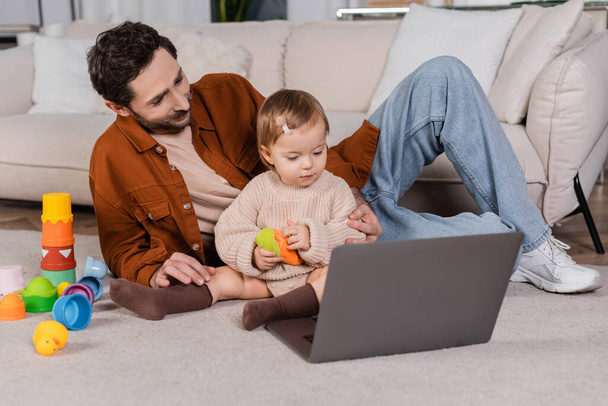 Father sitting near baby daughter with toy and laptop at home  - Foto, Imagem