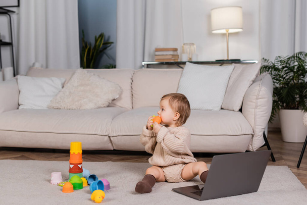 Vue latérale de bébé fille tenant jouet près d'un ordinateur portable dans le salon  - Photo, image