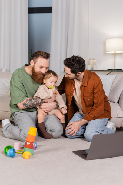 Gay couple hugging daughter near toys and laptop at home  - Photo, Image