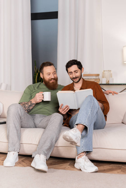 Positive gay couple with coffee reading book at home  - Fotoğraf, Görsel