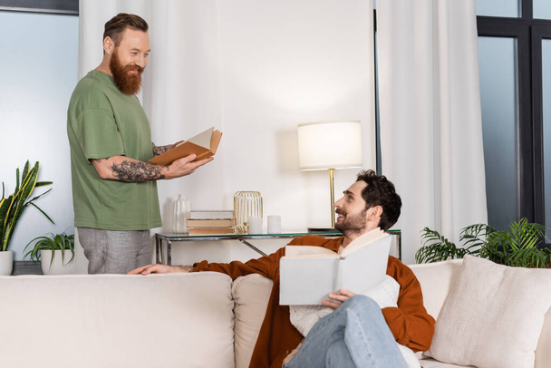 Smiling same sex couple holding books in living room  - Photo, Image